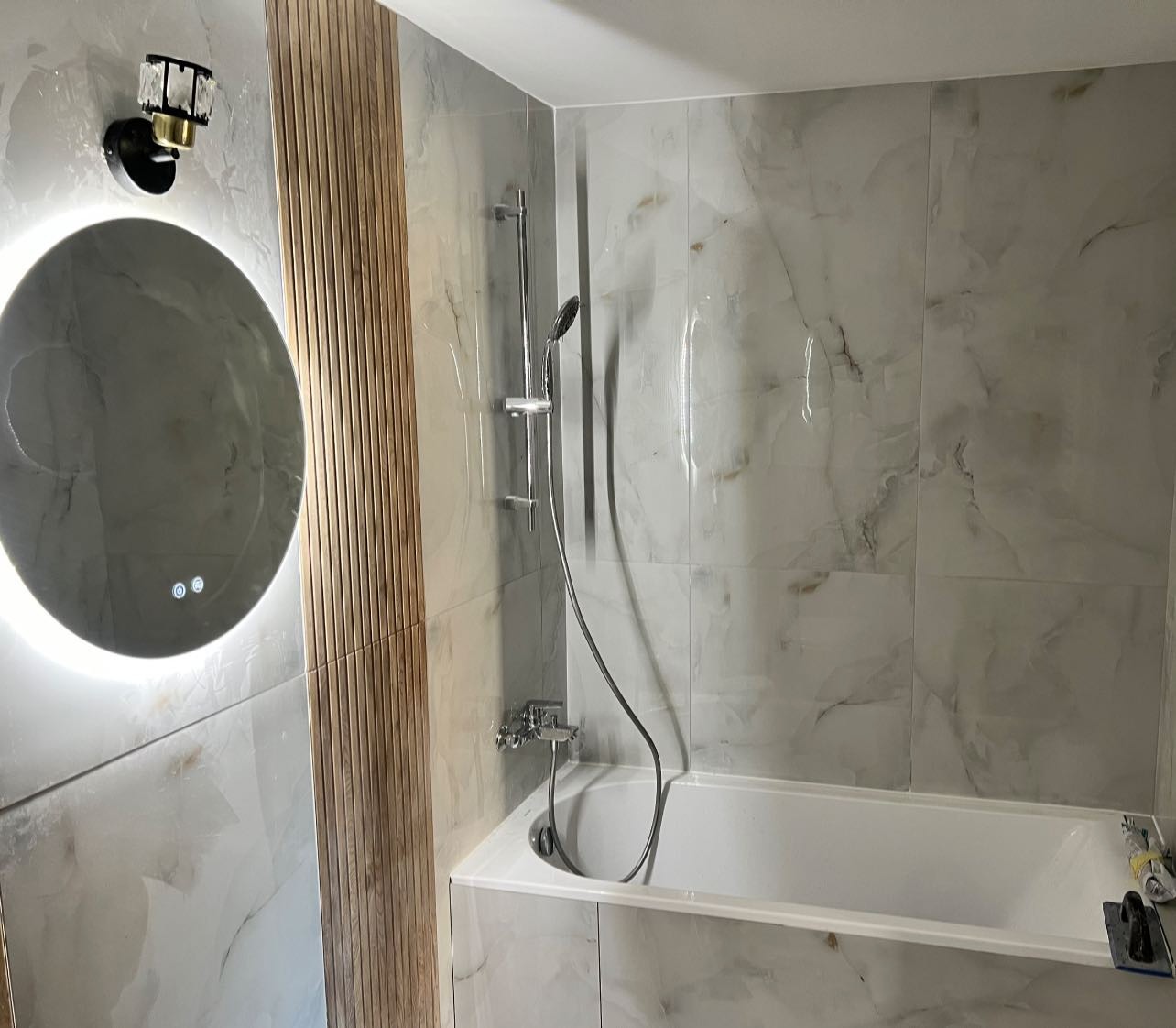 Wykończona łazienka z wanną, marmurowymi płytkami i okrągłym lustrem z podświetleniem LED. Dekoracyjny panel drewniany dodaje ciepła. Widoczne narzędzia glazurnicze.
