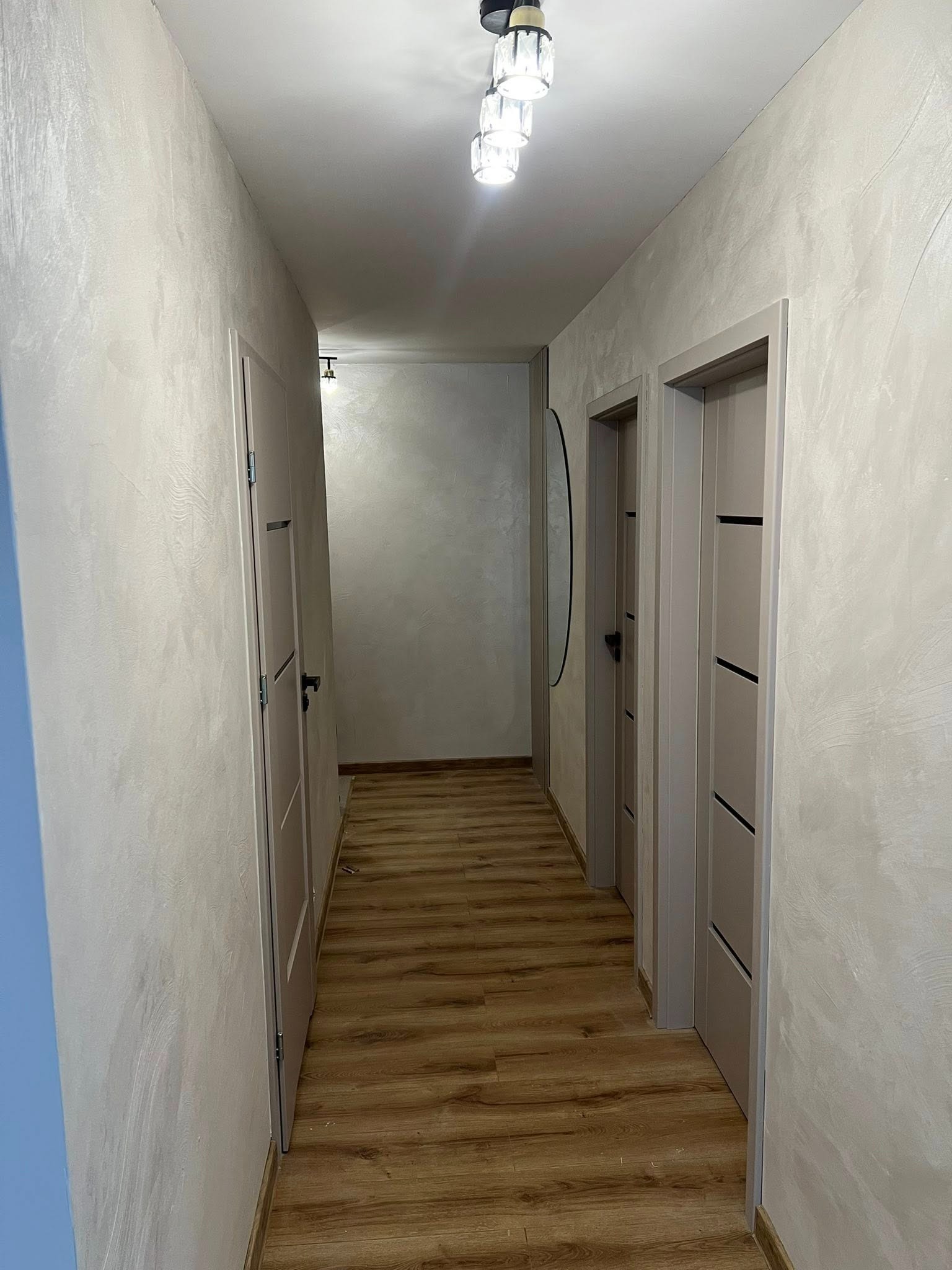 Wykończony korytarz z drewnianą podłogą, teksturowanymi ścianami w odcieniach beżu i szarości oraz nowoczesnym oświetleniem sufitowym. Widoczne drzwi z minimalistycznym designem.