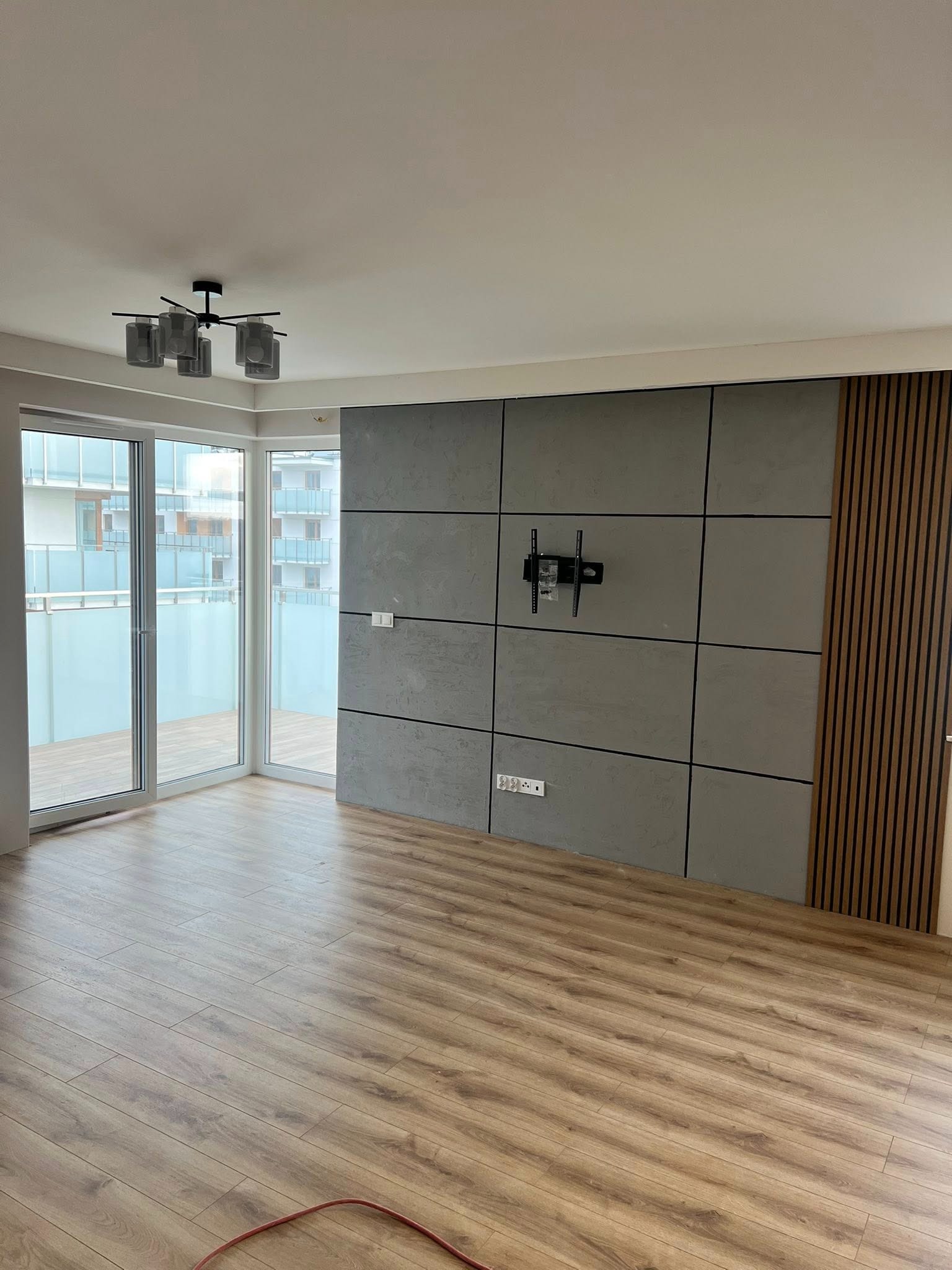 Nowoczesne wykończenie wnętrza: panele ścienne imitujące beton, drewniane lamele, parkiet. Widoczny uchwyt na TV i balkon z widokiem na budynek.
