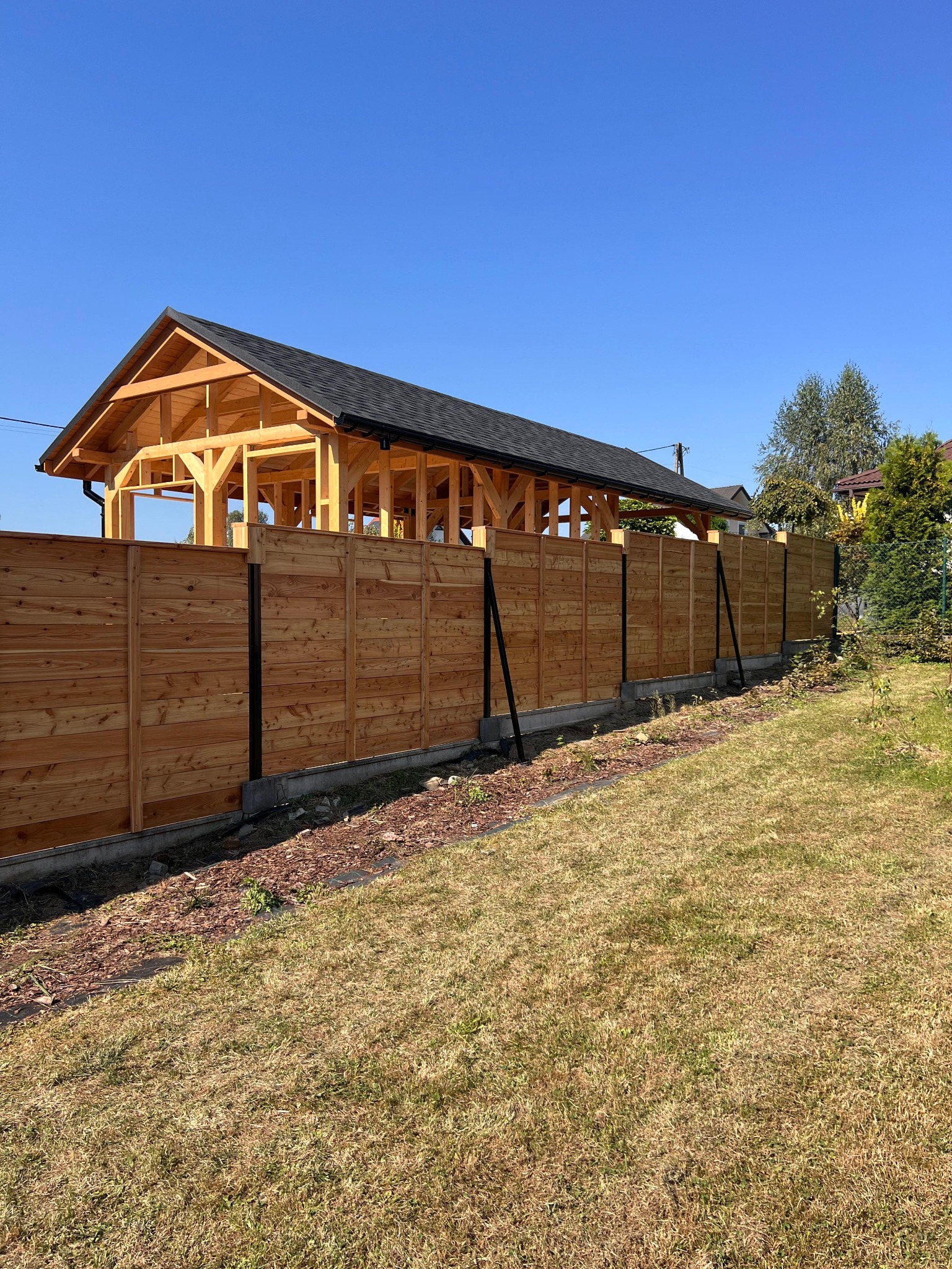 Nowoczesne ogrodzenie z drewnianych paneli, z widoczną konstrukcją altany w tle, na tle błękitnego nieba. Elementy metalowe w kolorze czarnym.