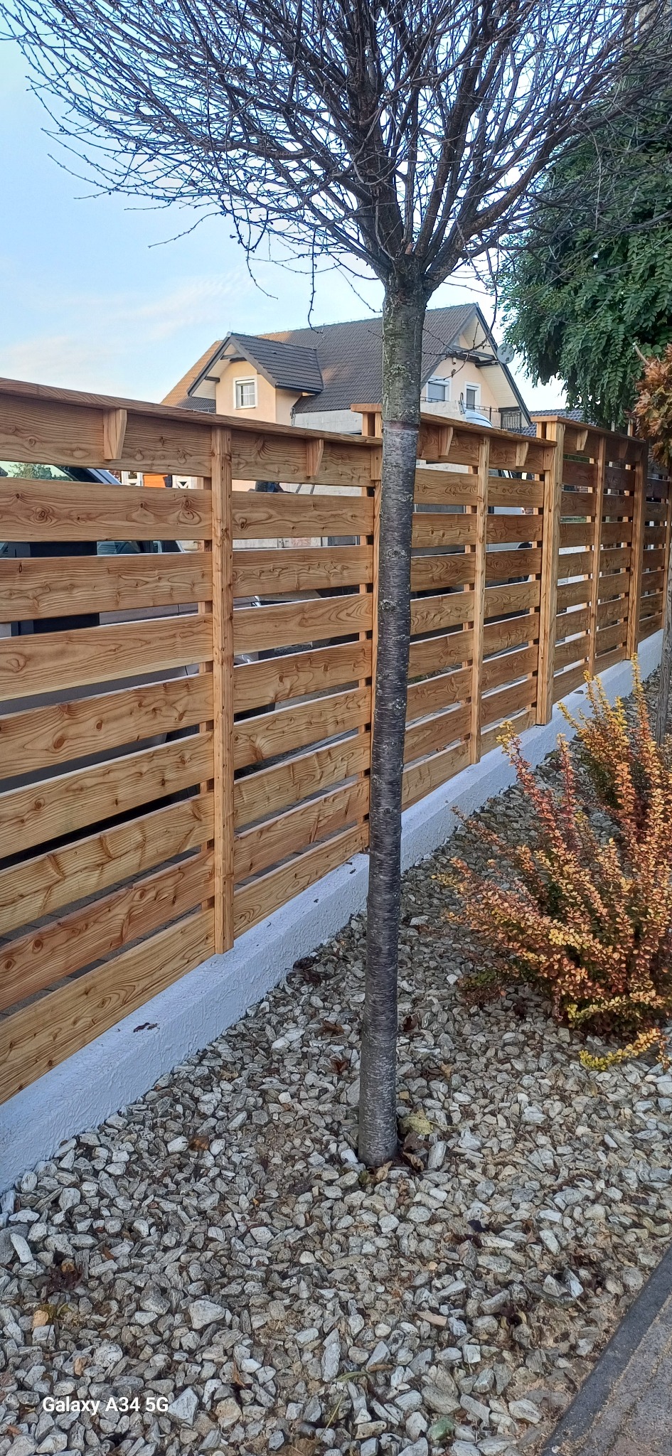 Poziome, drewniane panele ogrodzeniowe z widocznym drzewem na tle domu. Jasne drewno, szare kamienie i krzewy tworzą naturalny wygląd.