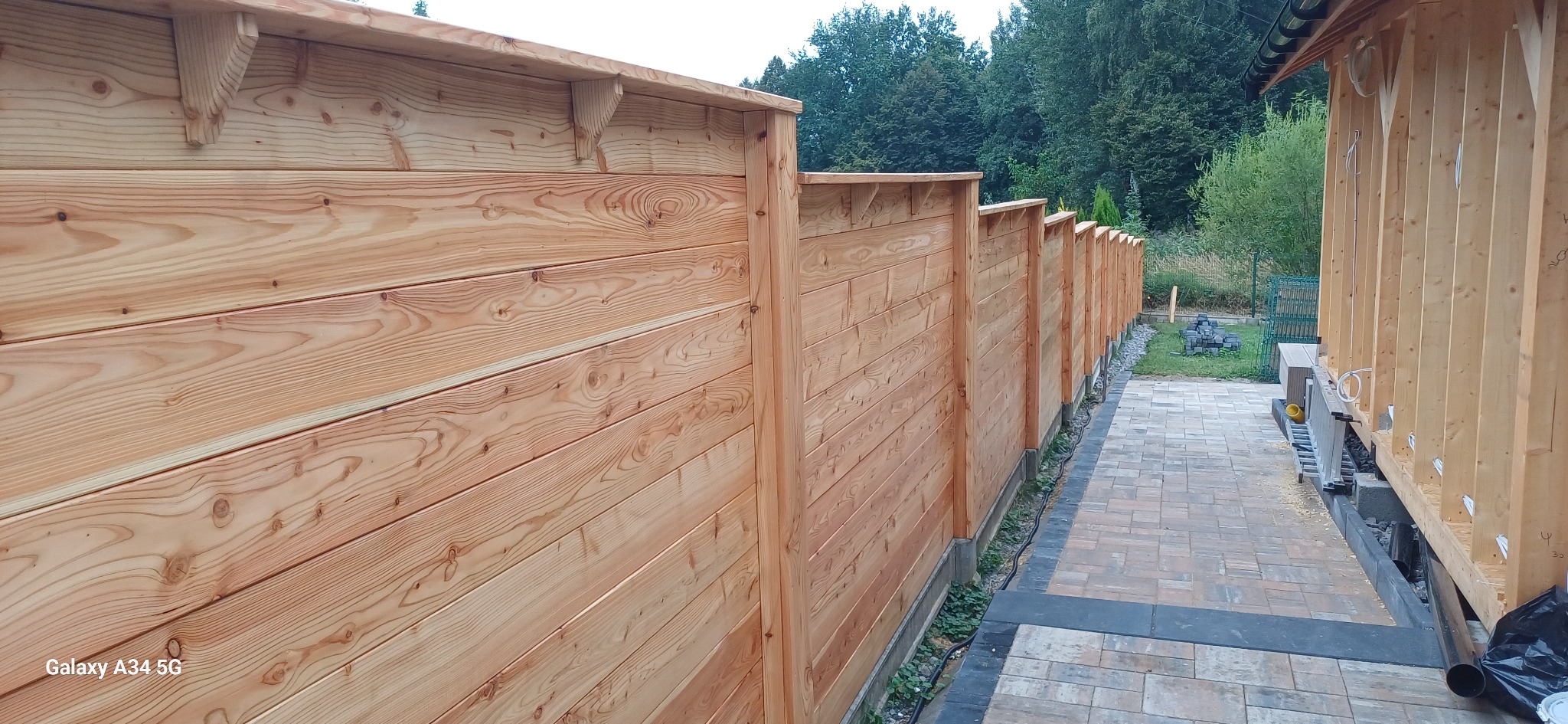 Długa, drewniana konstrukcja ogrodzeniowa z jasnego drewna, z widocznymi detalami łączeń i wspornikami, ciągnąca się wzdłuż ścieżki z kostki brukowej.