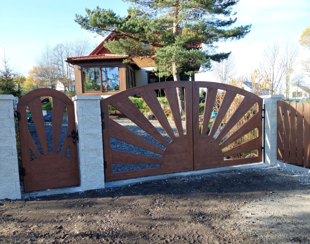 Brama wjazdowa i furtka z drewna, z motywem słońca, osadzone na kamiennych słupkach. W tle dom z brązowym dachem i zielone drzewa w słoneczny dzień.