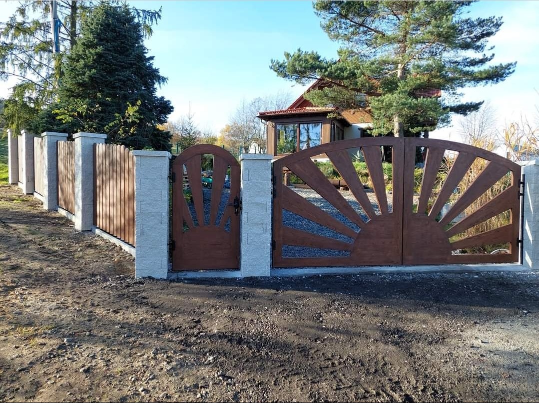 Nowoczesna brama wjazdowa i furtka z drewnianymi elementami w kształcie słońca, osadzone na słupkach z szarej kostki. W tle dom jednorodzinny i drzewa.