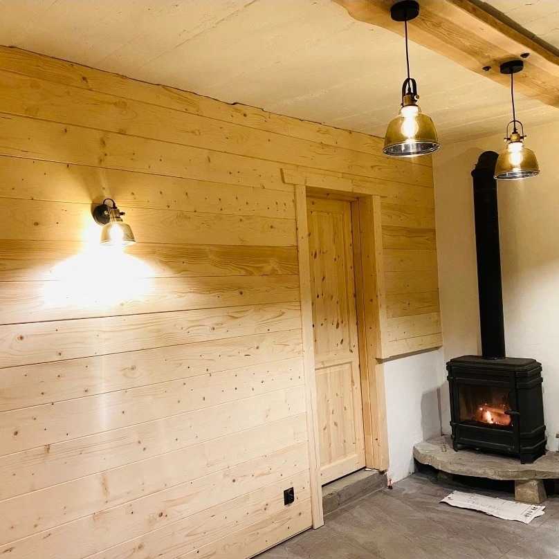 Drewniane wykończenie wnętrza z kominkiem w Mesznej. Ściany i drzwi z jasnego drewna, oświetlenie w stylu retro. Widoczny fragment sufitu z drewnianą belką.