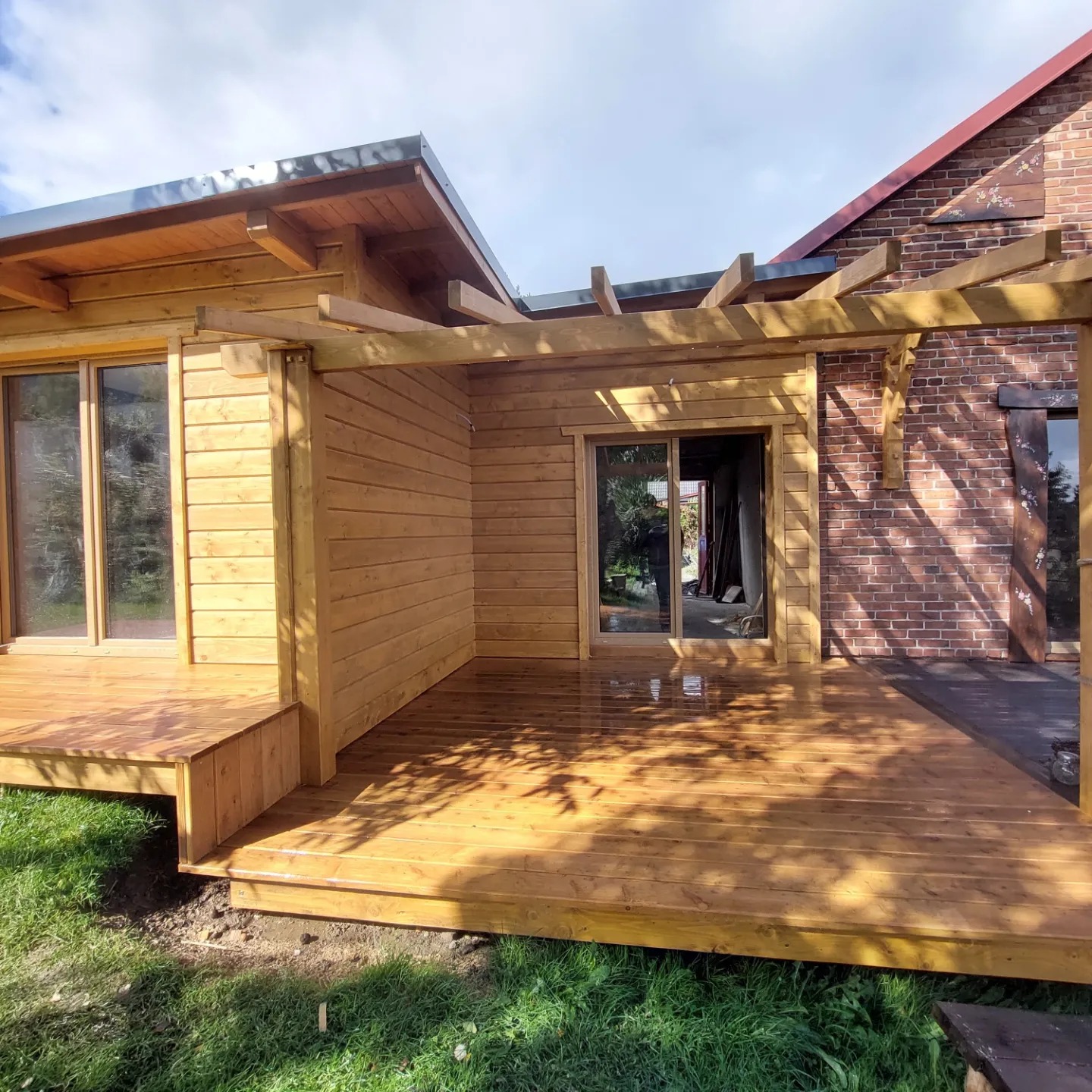 Drewniany taras przylegający do budynku z cegły i drewna, z elementami pergoli. Widoczne przesuwne drzwi i świeżo położona, lakierowana podłoga tarasu.