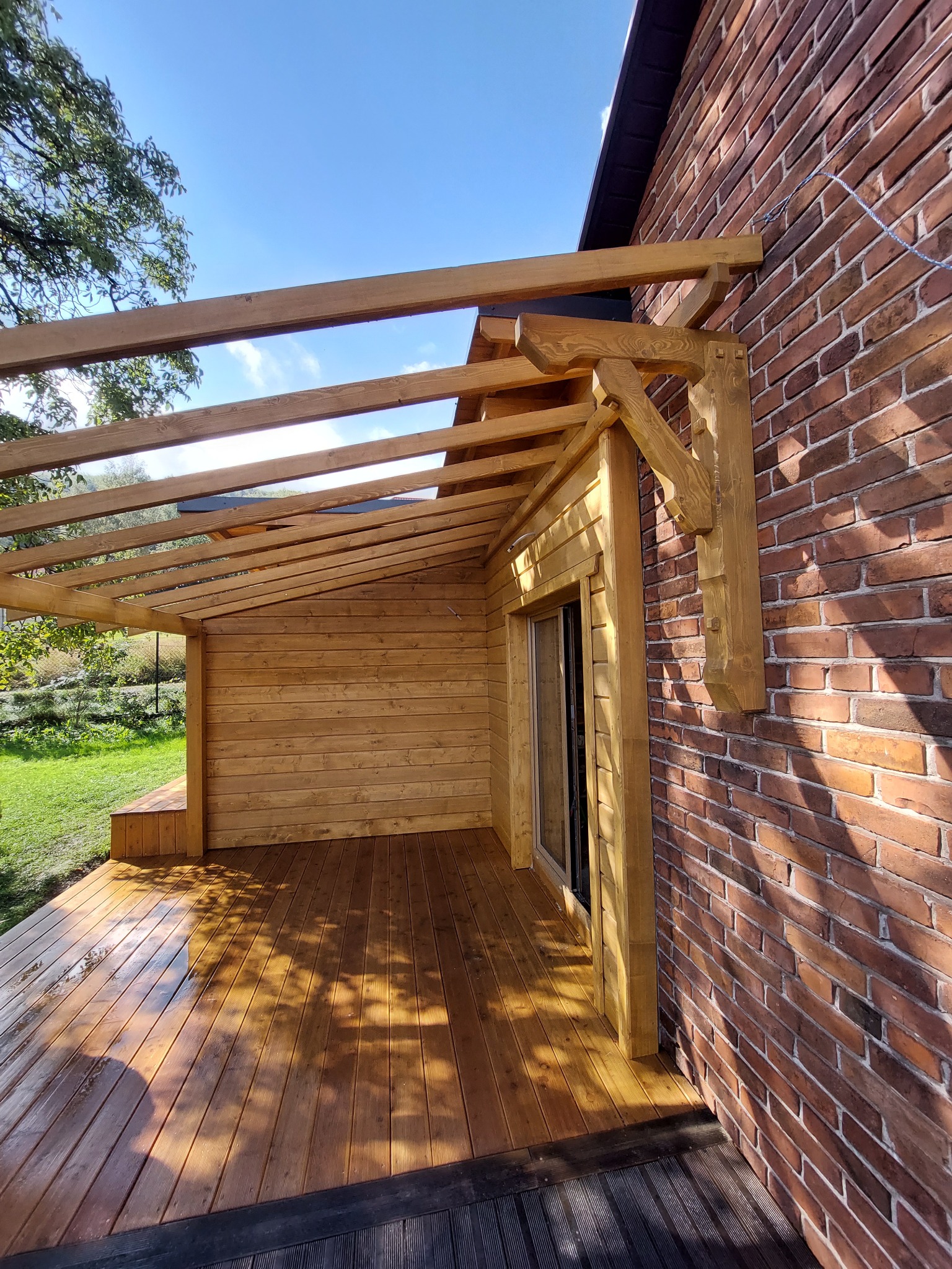 Drewniana konstrukcja tarasu przylegająca do ceglanego budynku, z widoczną podłogą z desek i elementami drewnianej zabudowy. Jasne drewno kontrastuje z czerwoną cegłą.