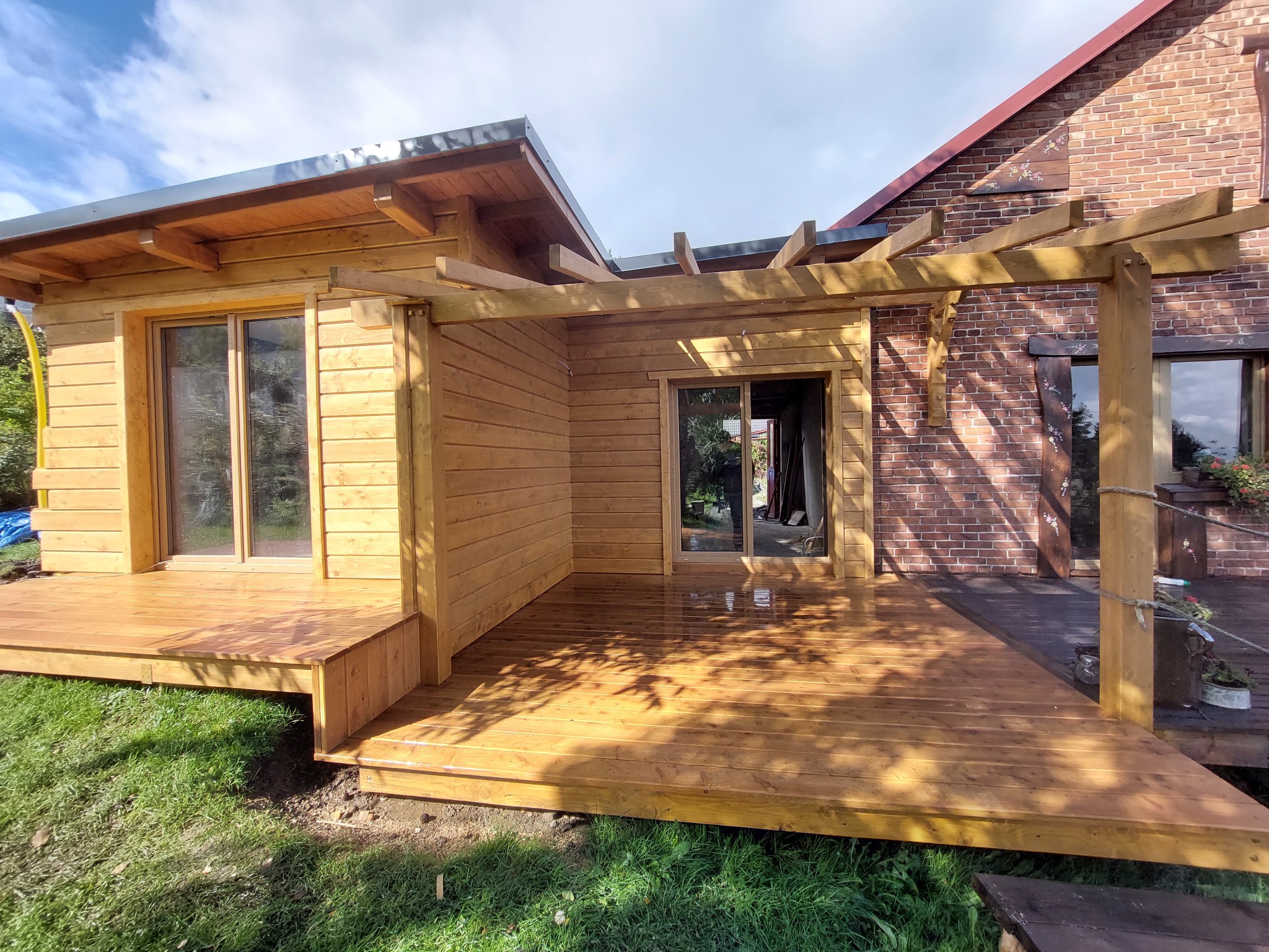 Drewniany taras z pergolą przy budynku z cegły, z widocznymi przeszkleniami i elementami dekoracyjnymi. Jasne drewno, trawa i częściowo zachmurzone niebo w tle.