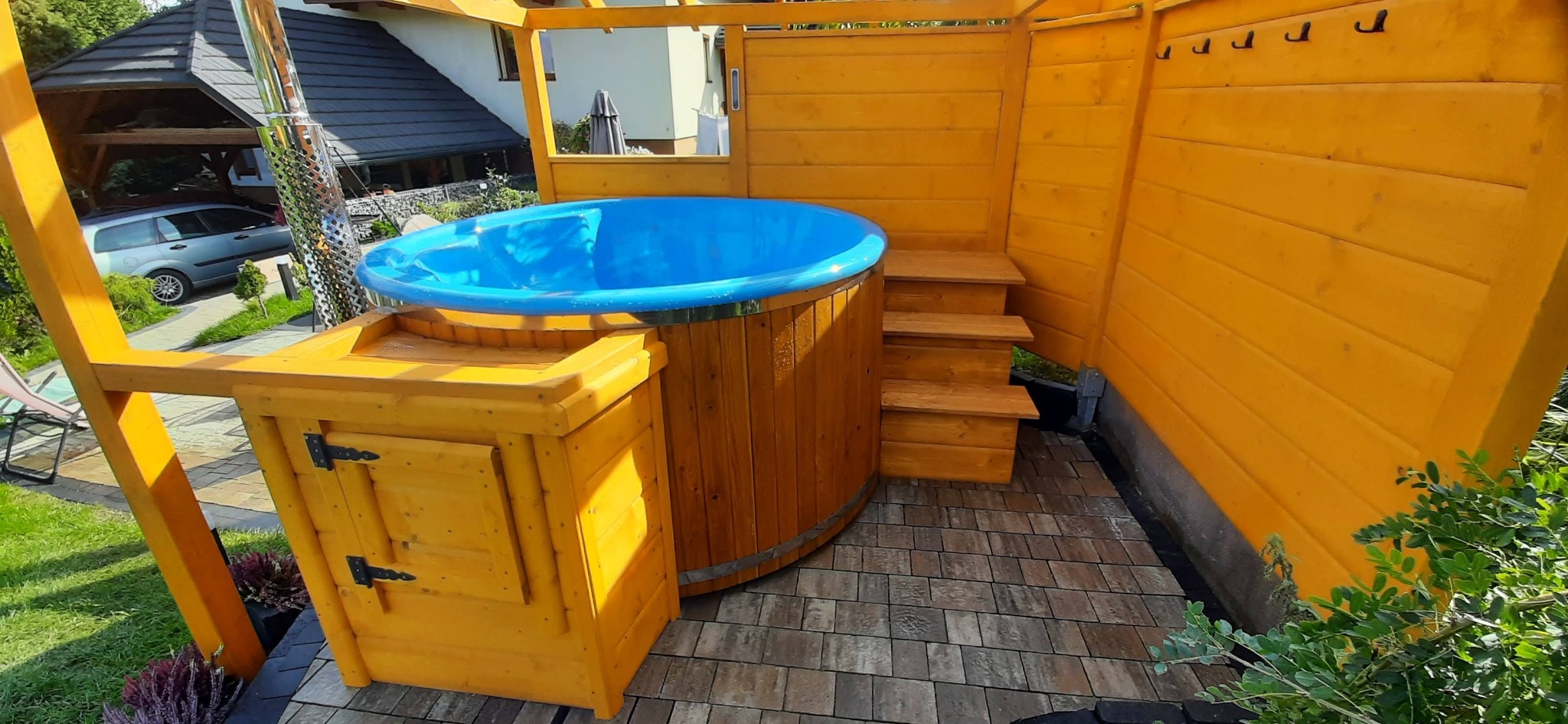 Drewniana balia ogrodowa z niebieskim wkładem, obudowana żółtą konstrukcją z drewna, z widocznymi schodkami i szafką na akcesoria, na tle kostki brukowej i zieleni.