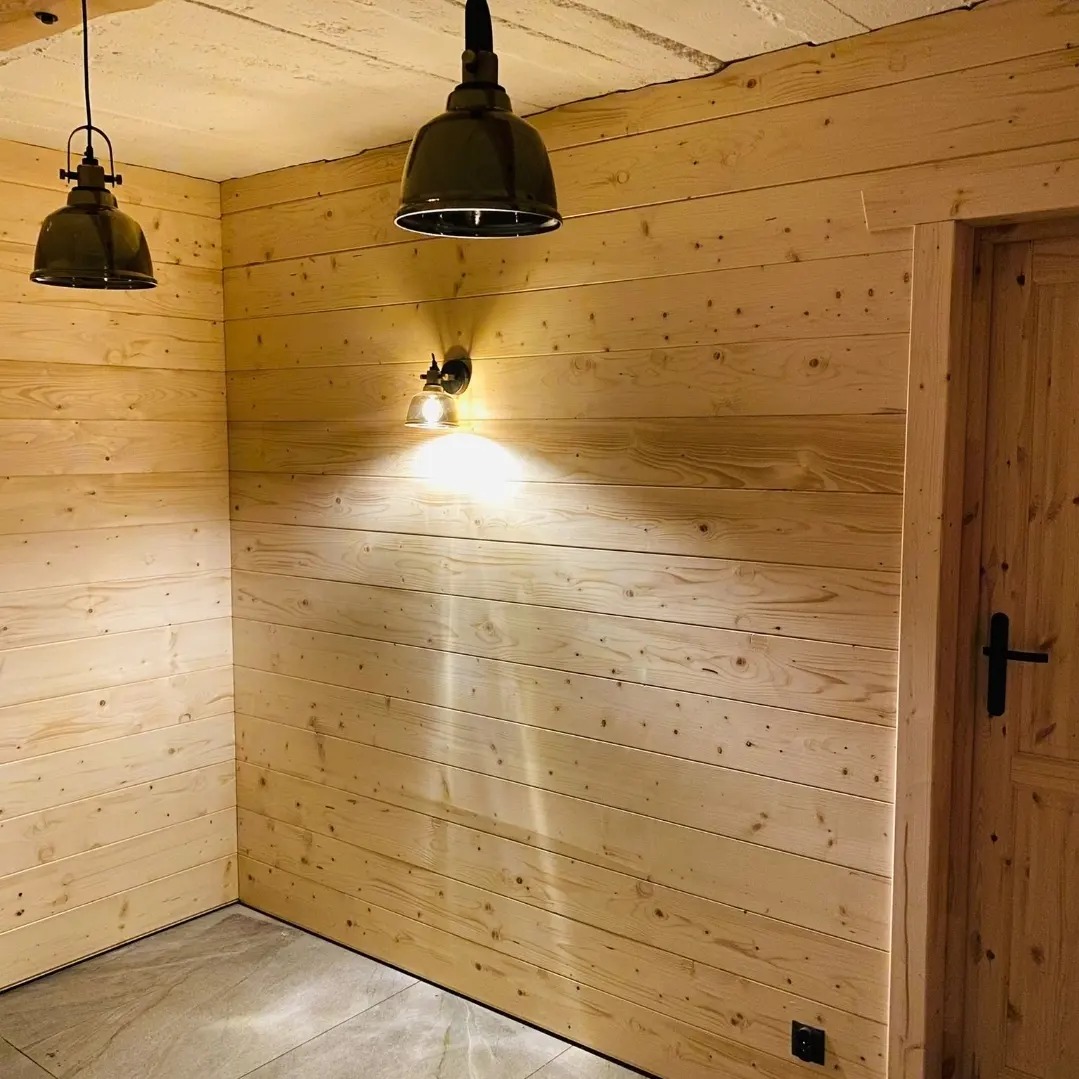 Drewniane panele ścienne w jasnym odcieniu, oświetlone lampami w stylu industrialnym, tworzą przytulne wnętrze z widoczną fakturą drewna i minimalistycznym wykończeniem.