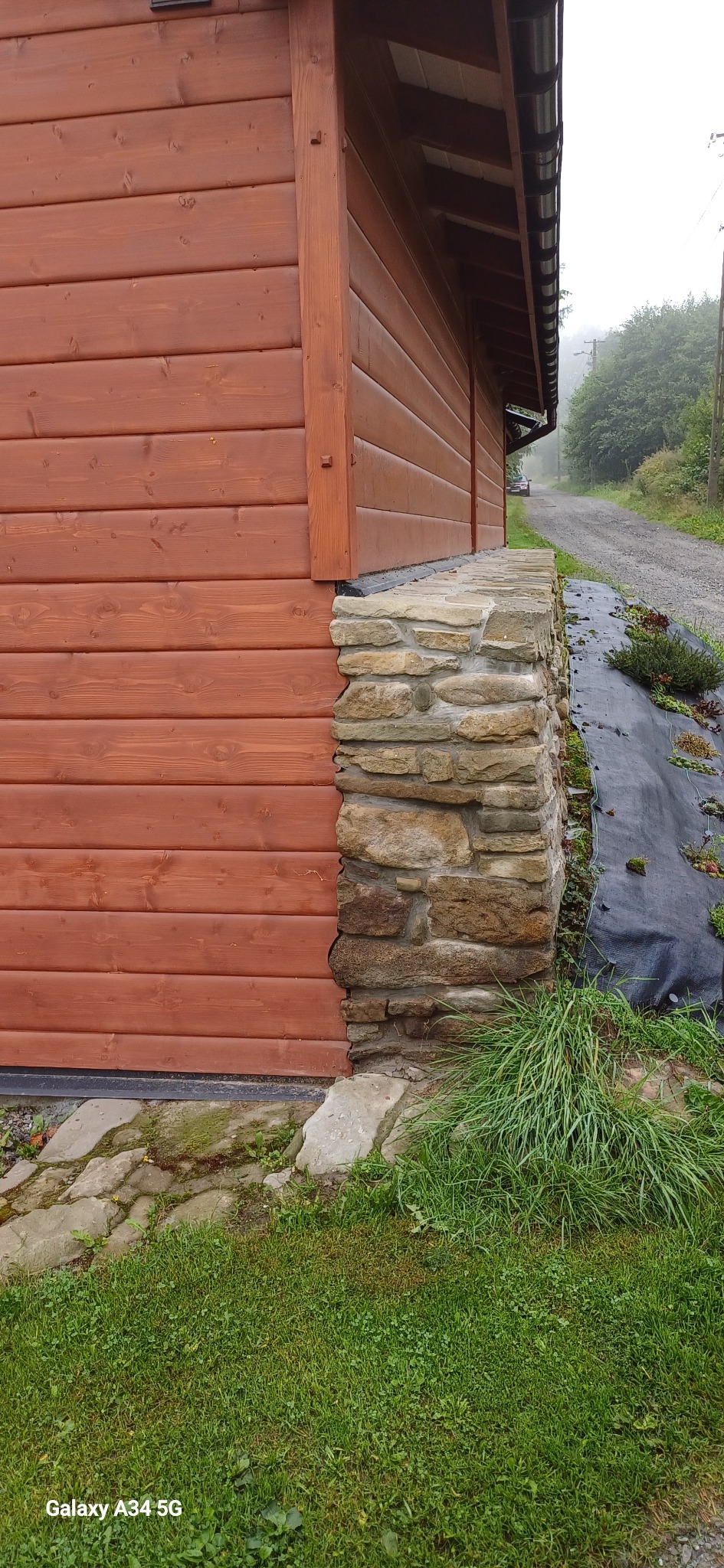 Fragment ściany budynku z drewnianą boazerią i kamienną podmurówką. Widoczny fragment dachu, trawa i droga w tle. Ujęcie z bliska, podkreślające detale wykończenia.