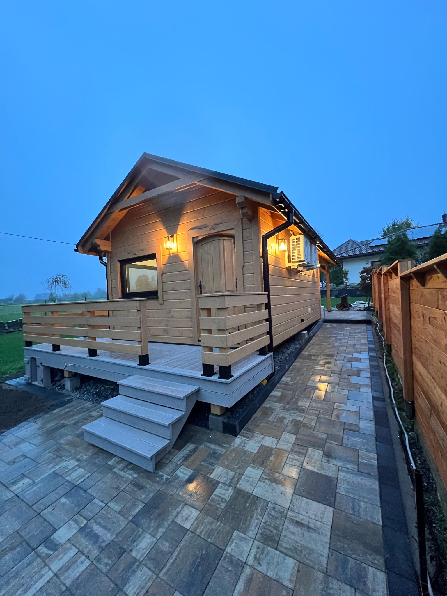 Drewniany domek letniskowy z tarasem i balustradą, oświetlony lampami, z klimatyzatorem na ścianie, otoczony kostką brukową i drewnianym ogrodzeniem w wieczornym świetle.