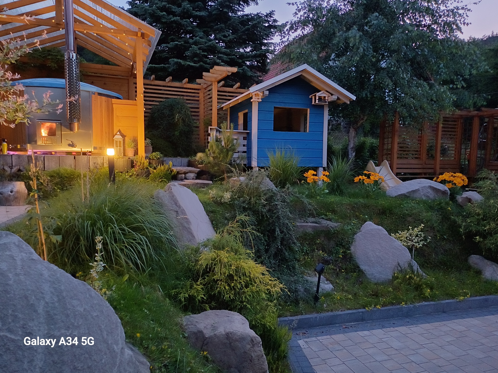 Ogród z drewnianą altaną dla dzieci, jacuzzi pod zadaszeniem i pergolą. Kamienne elementy dekoracyjne i bujna roślinność tworzą unikalny krajobraz.