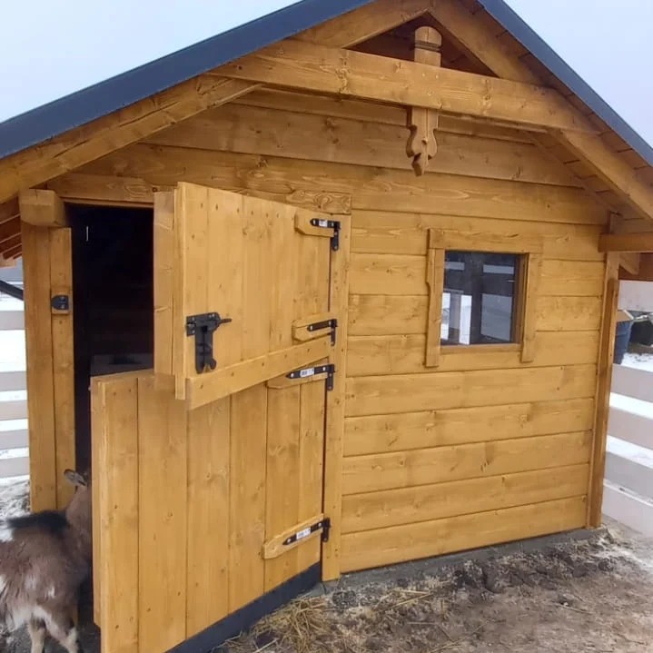 Drewniana konstrukcja z otwartymi drzwiami, prawdopodobnie mała stajnia lub domek gospodarczy. Widoczna koza wchodząca do środka. Całość w naturalnym odcieniu drewna.