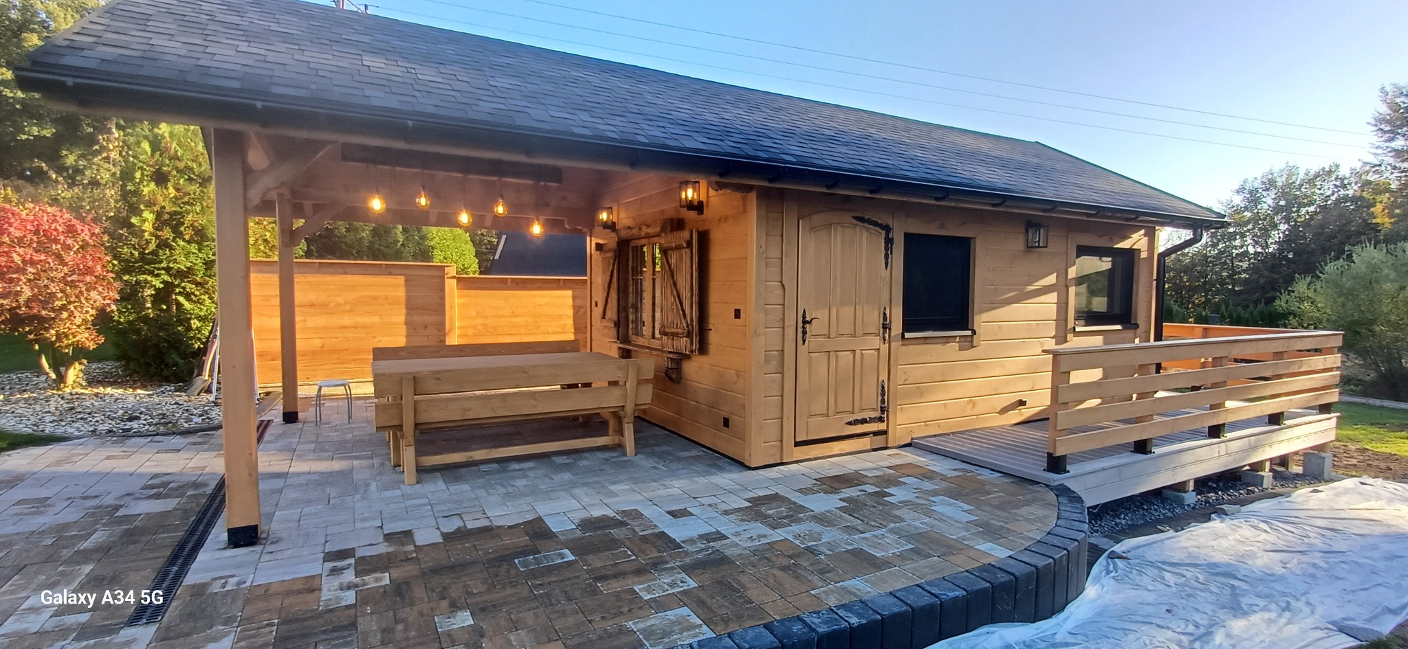 Drewniana altana z oświetleniem i stołem na wybrukowanym tarasie. Widoczne drewniane elementy konstrukcyjne i dekoracyjne. Całość w ciepłej kolorystyce.