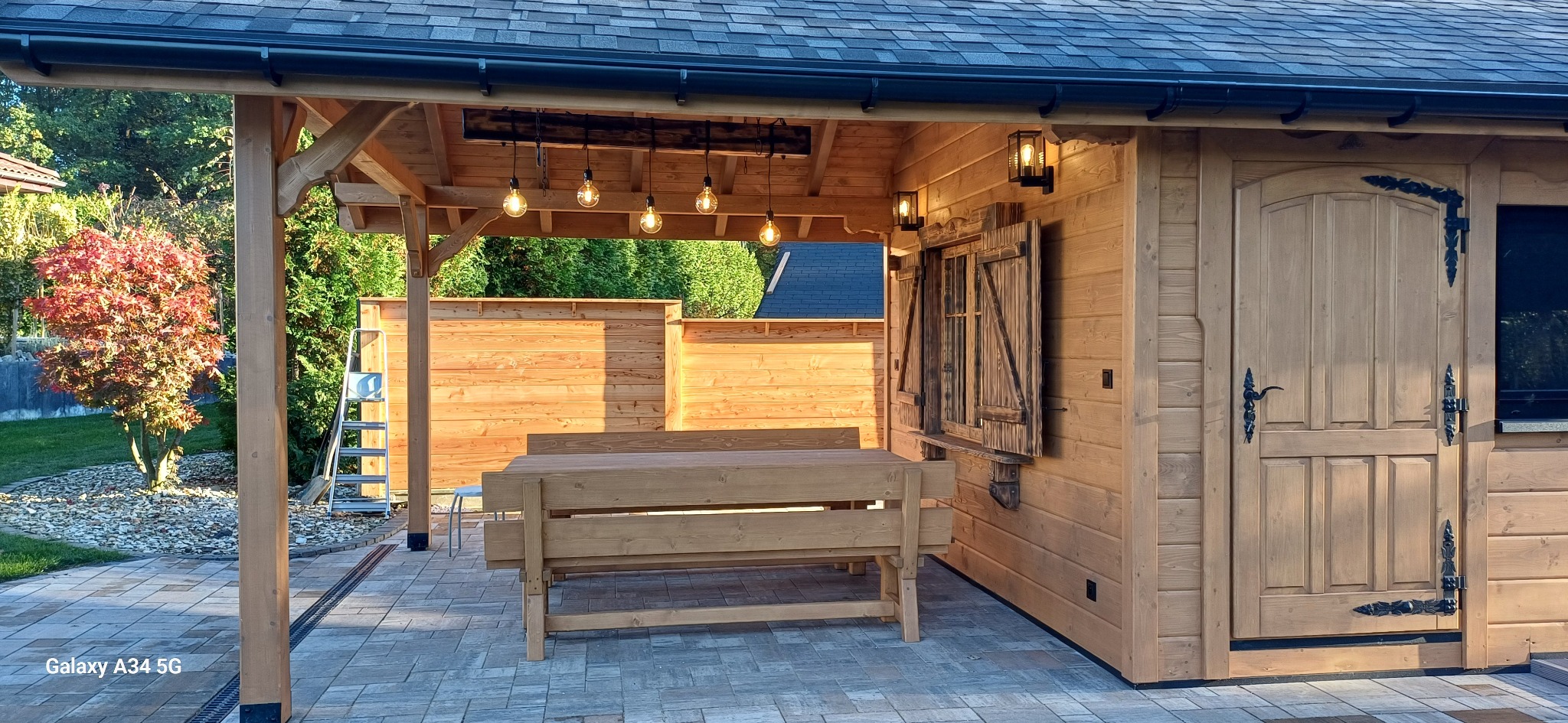 Drewniana konstrukcja z oświetleniem i stołem na zewnątrz, z elementami dekoracyjnymi i roślinnością w tle. Widoczna drabina oparta o ścianę.