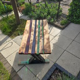 Przemysław Helpa - Stolik z drewnianym blatem i żywicą epoksydową w kolorach: brązowym, fioletowym i zielonym. Metalowe nogi. Ustawiony na tarasie z kostki brukowej.