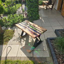 Przemysław Helpa - Drewniany stolik kawowy z żywicą epoksydową w kolorach tęczy, stojący na tarasie z kostki brukowej w słoneczny dzień. W tle zieleń ogrodu i osoba.