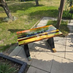 Przemysław Helpa - Kolorowy stolik kawowy z żywicy epoksydowej na tarasie z kostki brukowej. W tle trawnik z trampoliną i bawiącym się dzieckiem. Unikalny design i ręczne wykonanie.