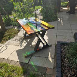 Przemysław Helpa - Kolorowy stolik kawowy z żywicy epoksydowej na tarasie, z unikalnym wzorem i metalowymi nogami. Widoczne refleksy światła na powierzchni blatu.