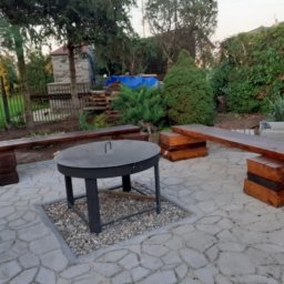 Przemysław Helpa - Rustykalny taras z drewnianymi ławkami i paleniskiem na kamiennej nawierzchni. W tle zieleń ogrodu i budynek gospodarczy.