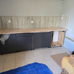 Przemysław Helpa - Częściowo zamontowane, nowoczesne szafki kuchenne z ciemnymi frontami i jasnym blatem, zabezpieczonym folią. Widoczne gniazdka elektryczne na ścianie.