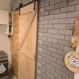 Przemysław Helpa - Drewniane drzwi przesuwne w stylu rustykalnym, zamontowane na szynie, oddzielają kuchnię od innego pomieszczenia. Szara ściana z cegły w tle.