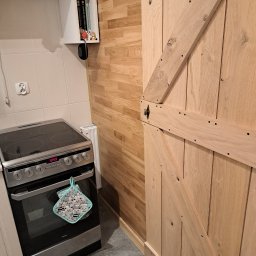 Przemysław Helpa - Kuchnia z drewnianą ścianą i drzwiami w stylu rustykalnym. Widoczny fragment kuchenki i szafki. Drewno o jasnym odcieniu, widoczne słoje.