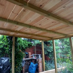 Przemysław Helpa - Drewniana konstrukcja zadaszenia tarasu w Obornikach, widok od dołu. Deski ułożone równolegle, zielone podpory, w tle roślinność i ceglany grill.