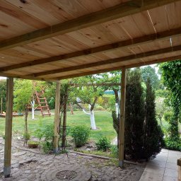 Przemysław Helpa - Zadaszenie tarasu z surowego drewna, widok na zielony ogród z elementami architektury ogrodowej i drzewami. Konstrukcja wsparta na drewnianych słupach.