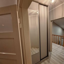 Szafa przesuwna z lustrem w przedpokoju, widok z boku otwartych drzwi. Podłoga wyłożona panelami, w tle schody z drewnianą balustradą.