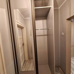 Wnętrze szafy przesuwnej z lustrem, widok na półki i drążek na ubrania. W lustrze odbija się fragment korytarza z drzwiami. Neutralna kolorystyka ścian i mebli.