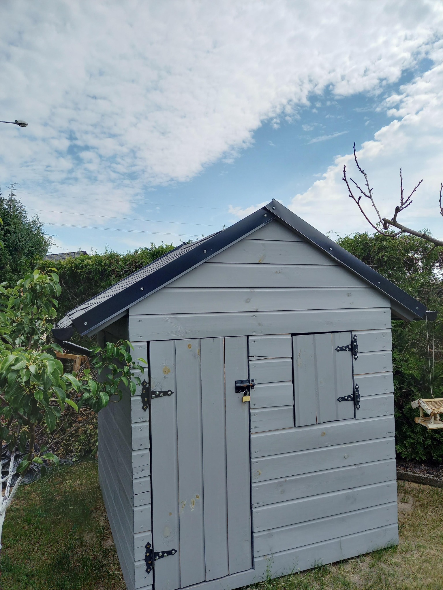 Szara, drewniana altanka ogrodowa z czarnym dachem i ozdobnymi zawiasami, zamknięta na kłódkę, otoczona zielenią. Widok z dołu, błękitne niebo w tle.