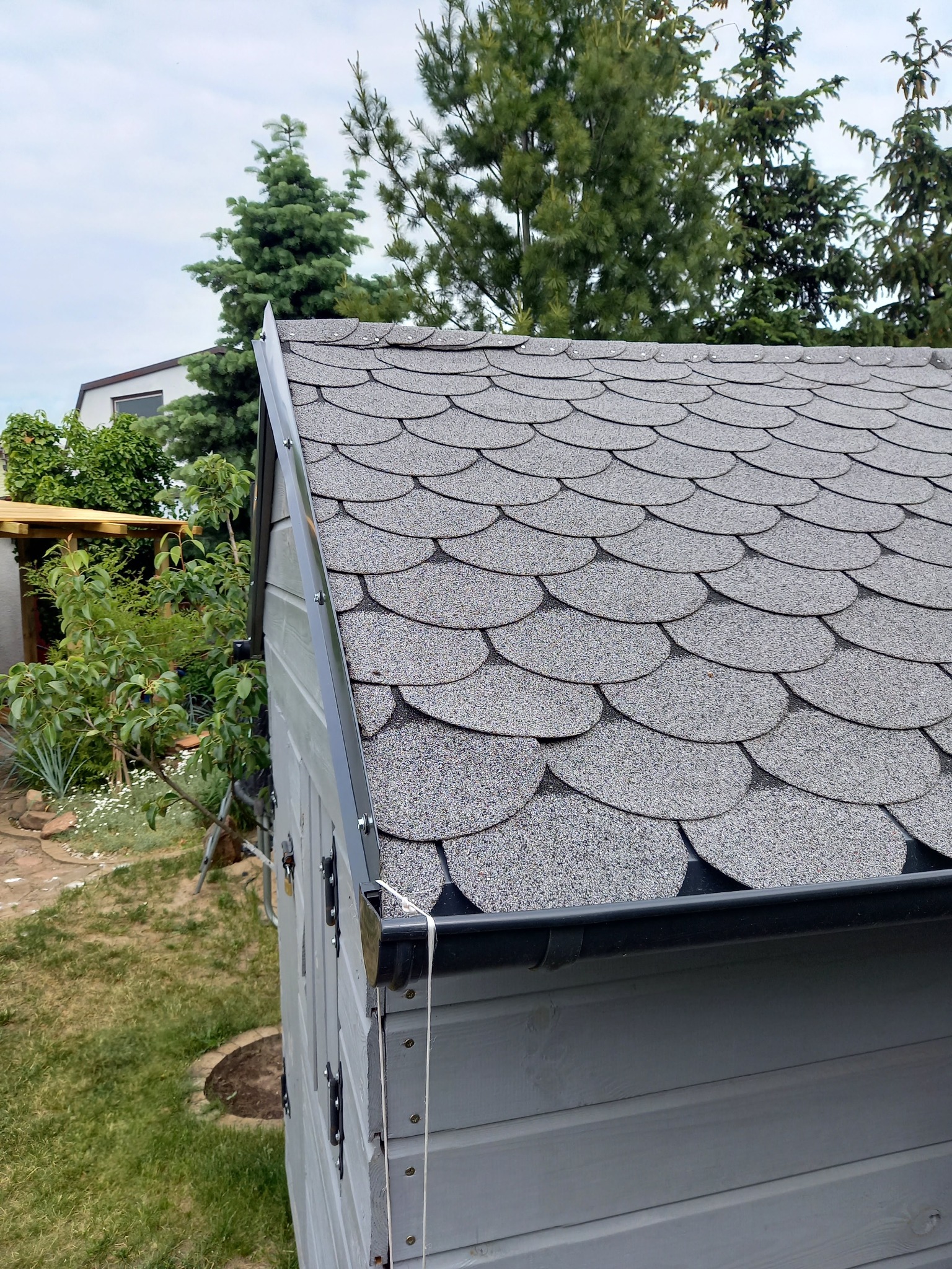 Szary dach pokryty gontem bitumicznym w kształcie rybiej łuski na drewnianej konstrukcji, z widocznymi elementami orynnowania i otoczeniem zieleni.