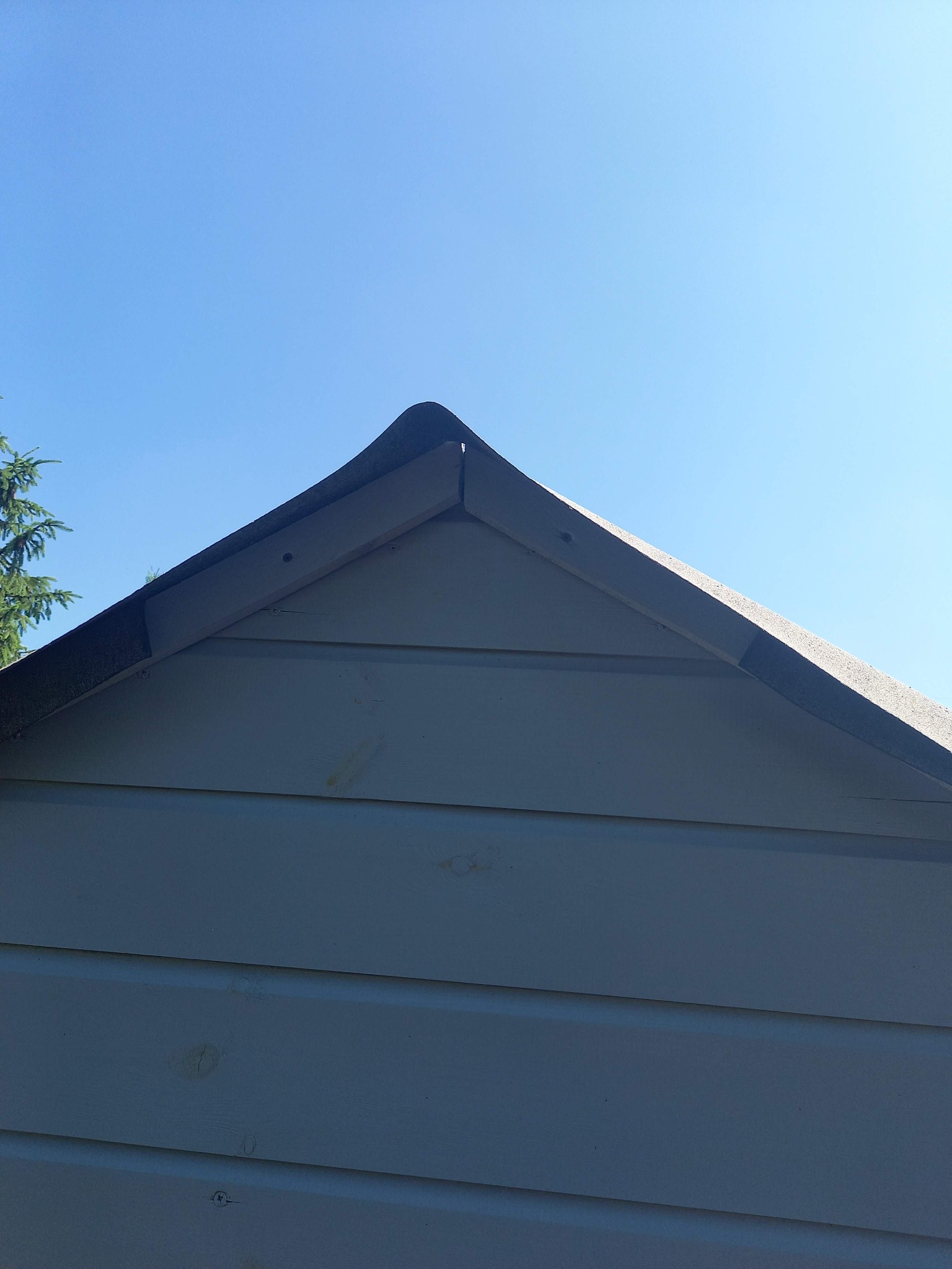 Fragment szczytu altany z drewnianą konstrukcją i pokryciem dachowym na tle błękitnego nieba. Widoczne deski elewacyjne i elementy wykończeniowe.