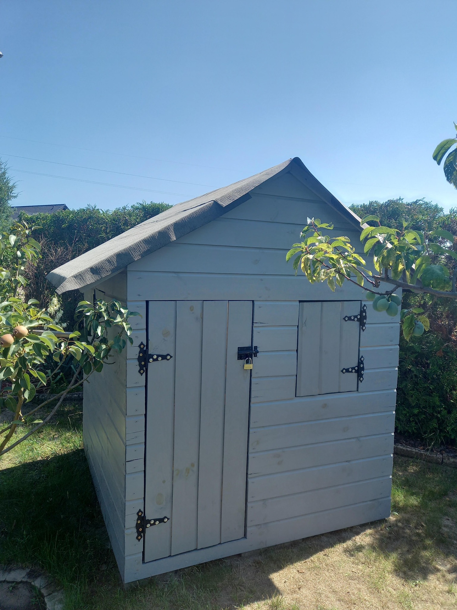 Drewniana, pomalowana na szaro altanka ogrodowa z zamkniętymi drzwiami i oknem, zabezpieczona kłódką, otoczona zielenią w słoneczny dzień.