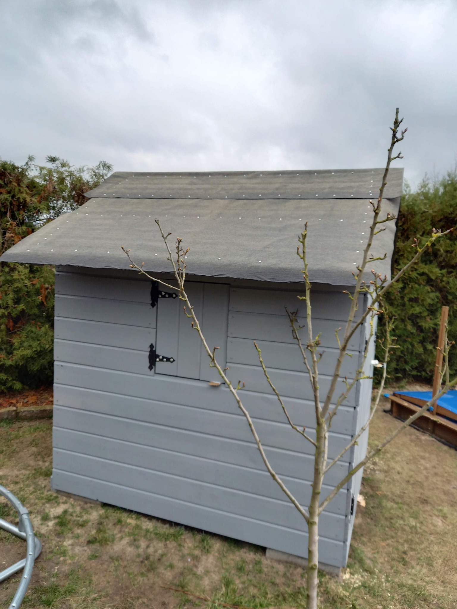 Szara, drewniana altanka ogrodowa z czarnymi zawiasami na tle pochmurnego nieba i zieleni. Ujęcie z dołu, z widocznym fragmentem metalowej konstrukcji.