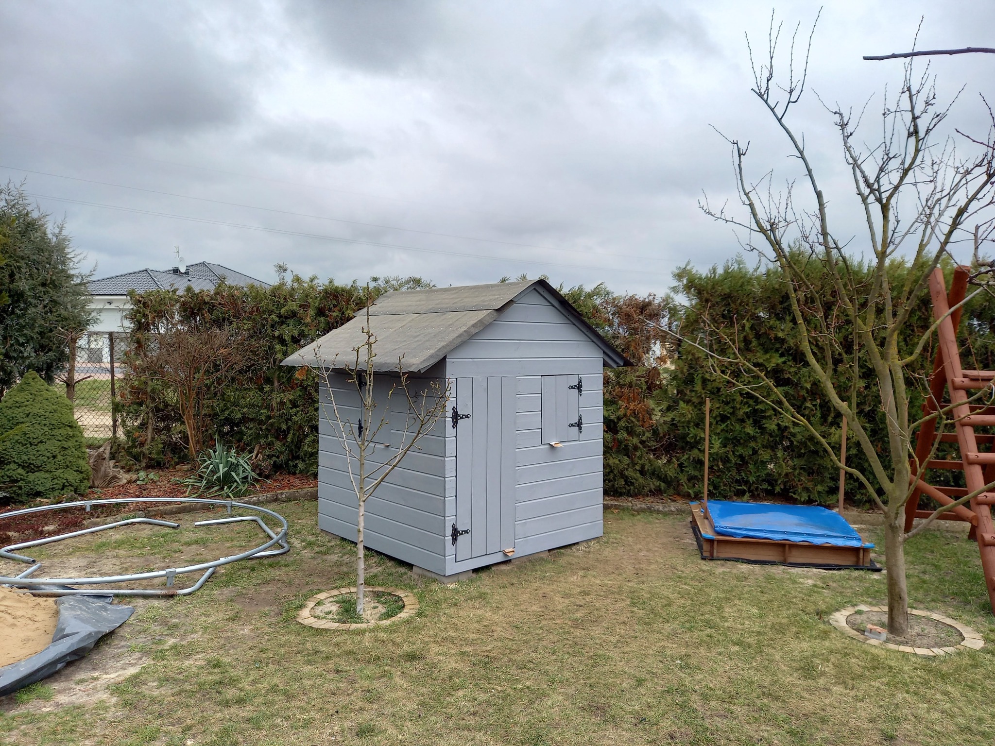 Szara, drewniana altanka narzędziowa z oknem i drzwiami na trawniku, obok piaskownicy z niebieskim przykryciem i rozłożonego trampoliny. Pochmurne niebo w tle.