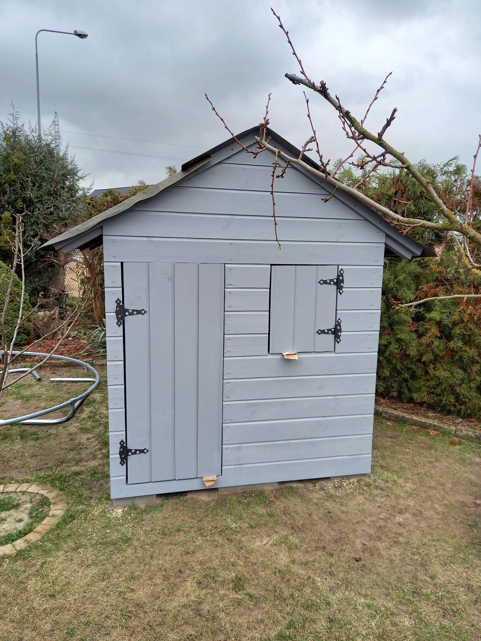 Szara, drewniana altana ogrodowa z czarnymi zawiasami, umieszczona na trawniku w ogrodzie. Widoczne drzewa i pochmurne niebo w tle.