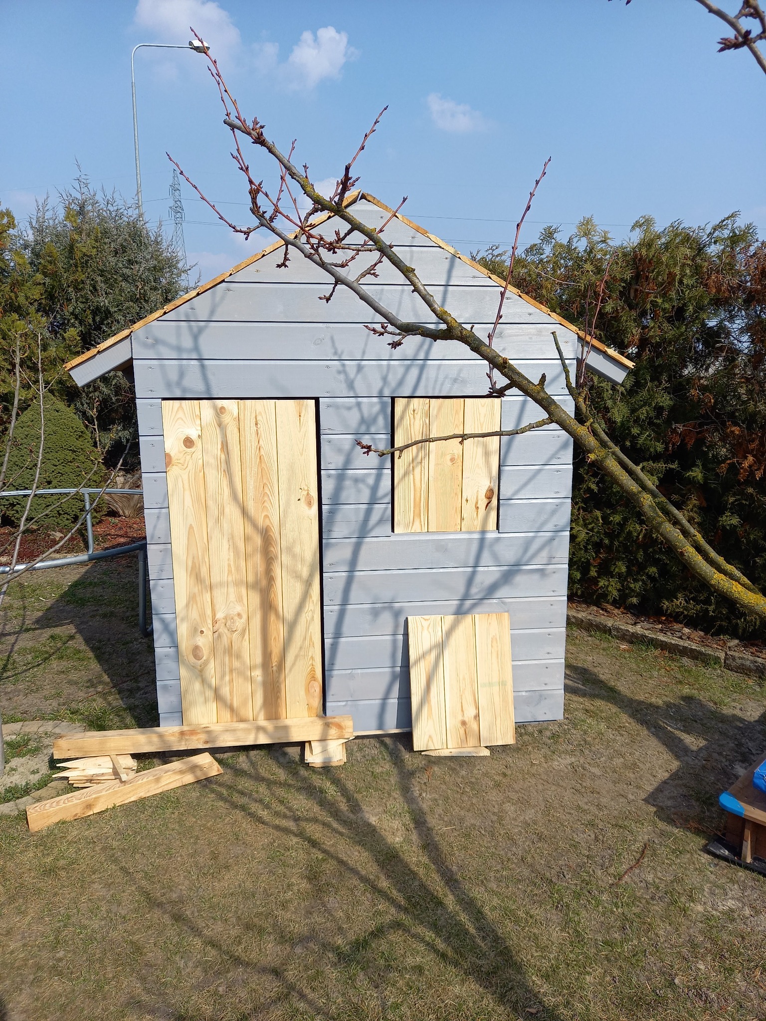 Szara, drewniana altanka w trakcie budowy, z surowymi, jasnymi drzwiami i okiennicą. Wokół trawa i drzewa, gałąź drzewa na pierwszym planie.