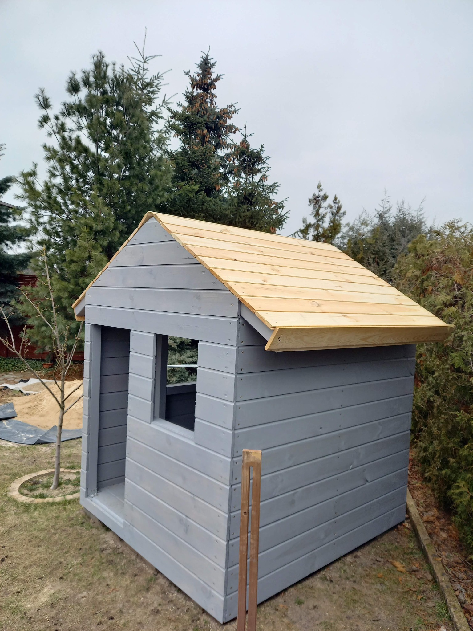 Szara drewniana altanka dla dzieci z jasnym dachem na trawniku, otoczona zielenią drzew i krzewów. Widoczne deski i konstrukcja. Element architektury ogrodowej.