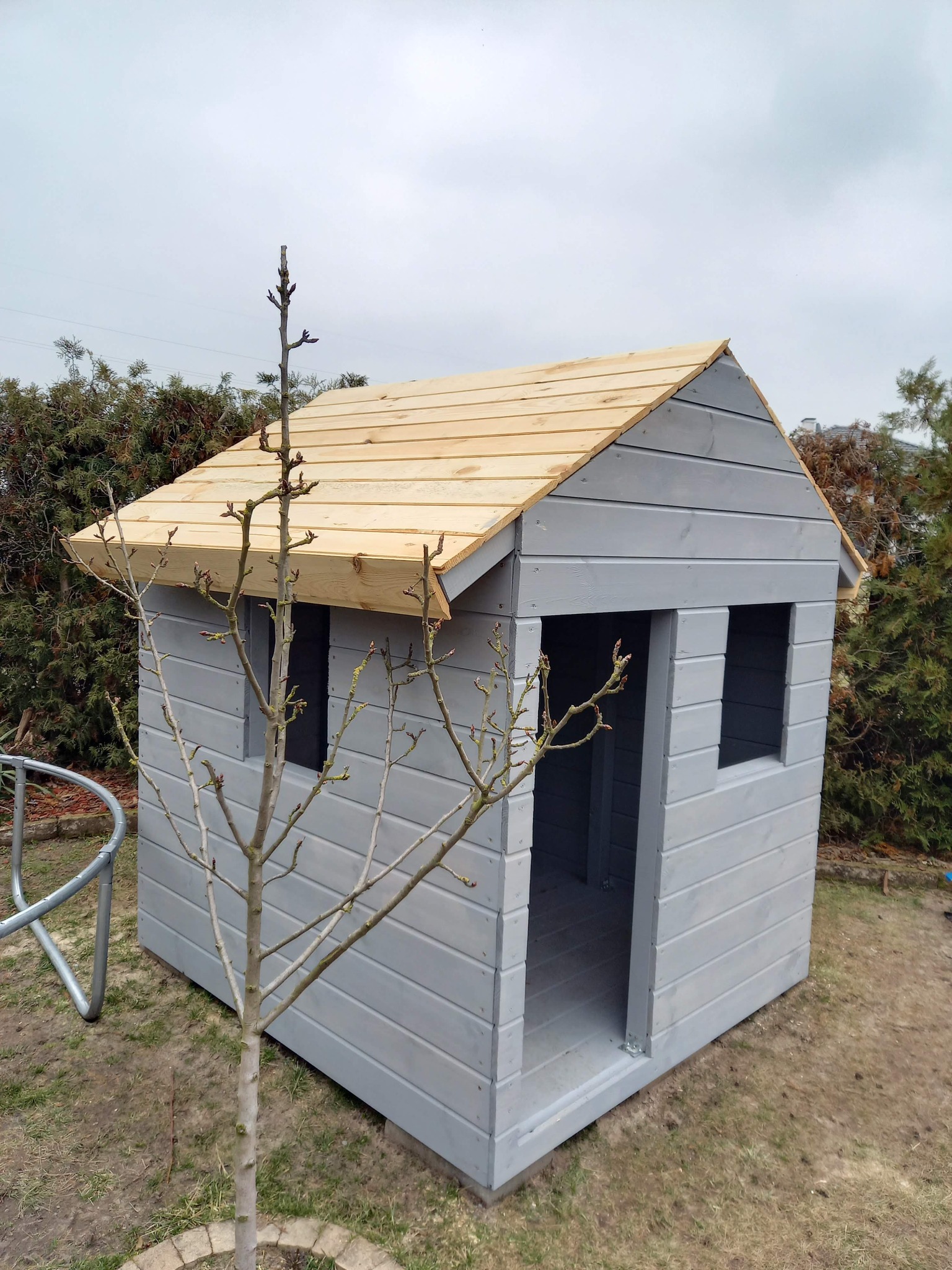 Szara, drewniana altanka dla dzieci z jasnym dachem, widziana z niskiej perspektywy, z młodym drzewkiem na pierwszym planie i zielonymi krzewami w tle.