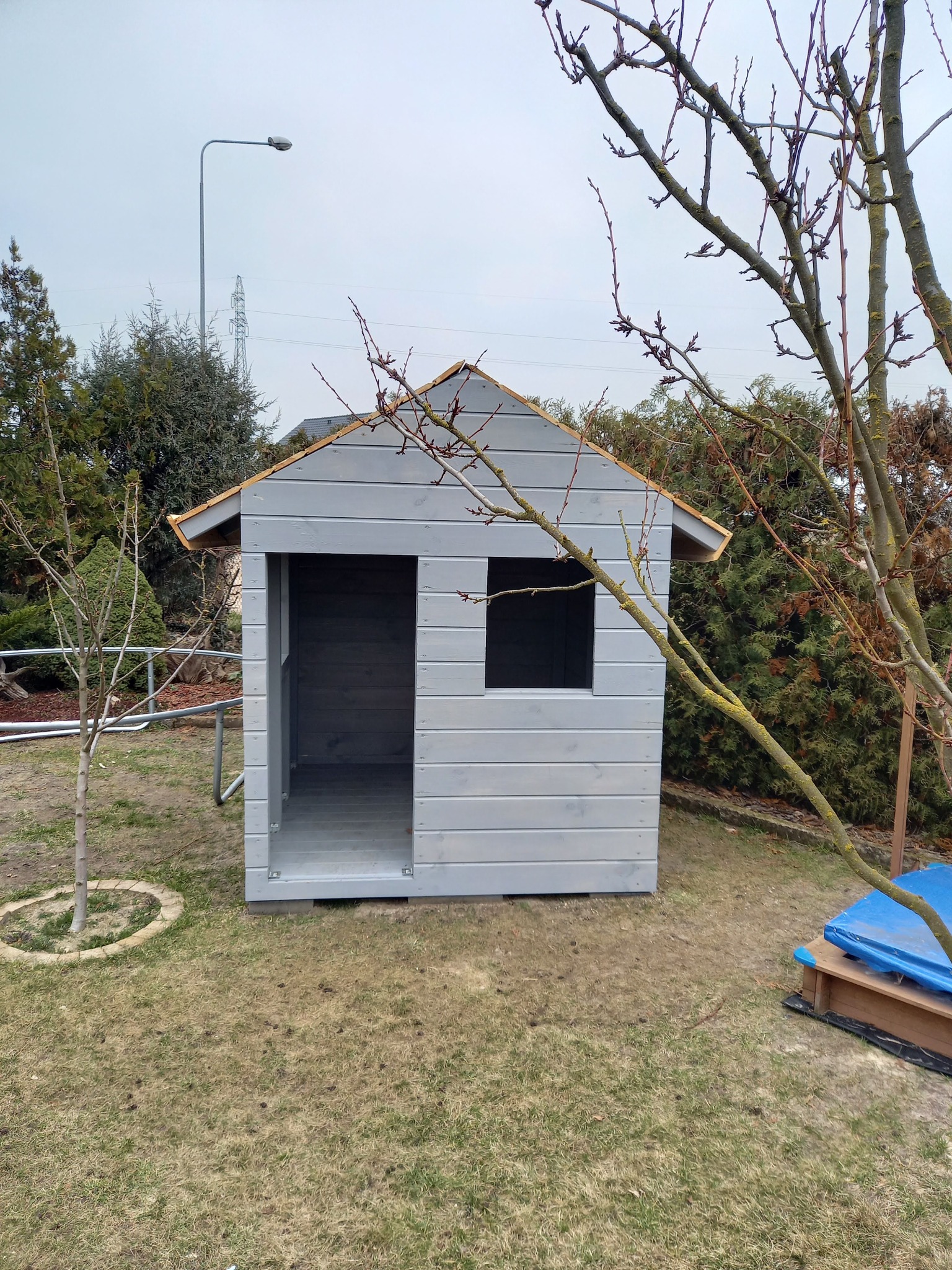 Szara, drewniana altanka dla dzieci z małym oknem, umieszczona na trawniku w ogrodzie. Widoczne drzewa i lampa uliczna w tle.