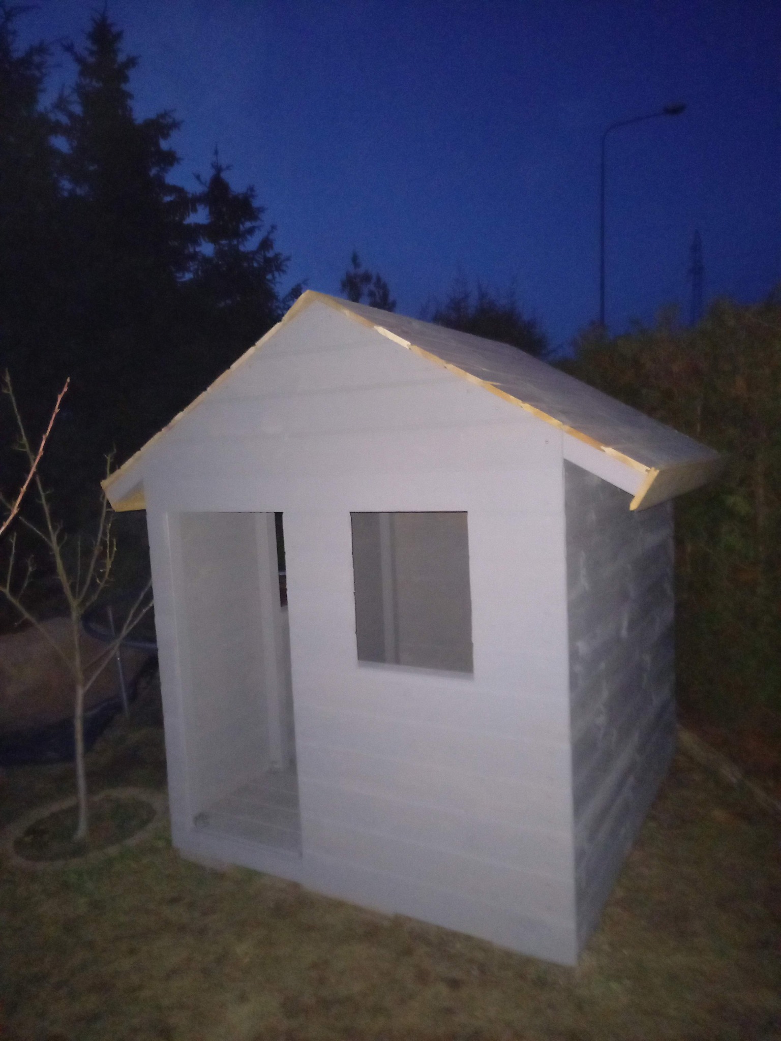 Mały, pomalowany na szaro domek dla dzieci z oknem i wejściem, stojący na trawie w ogrodzie, sfotografowany o zmierzchu.