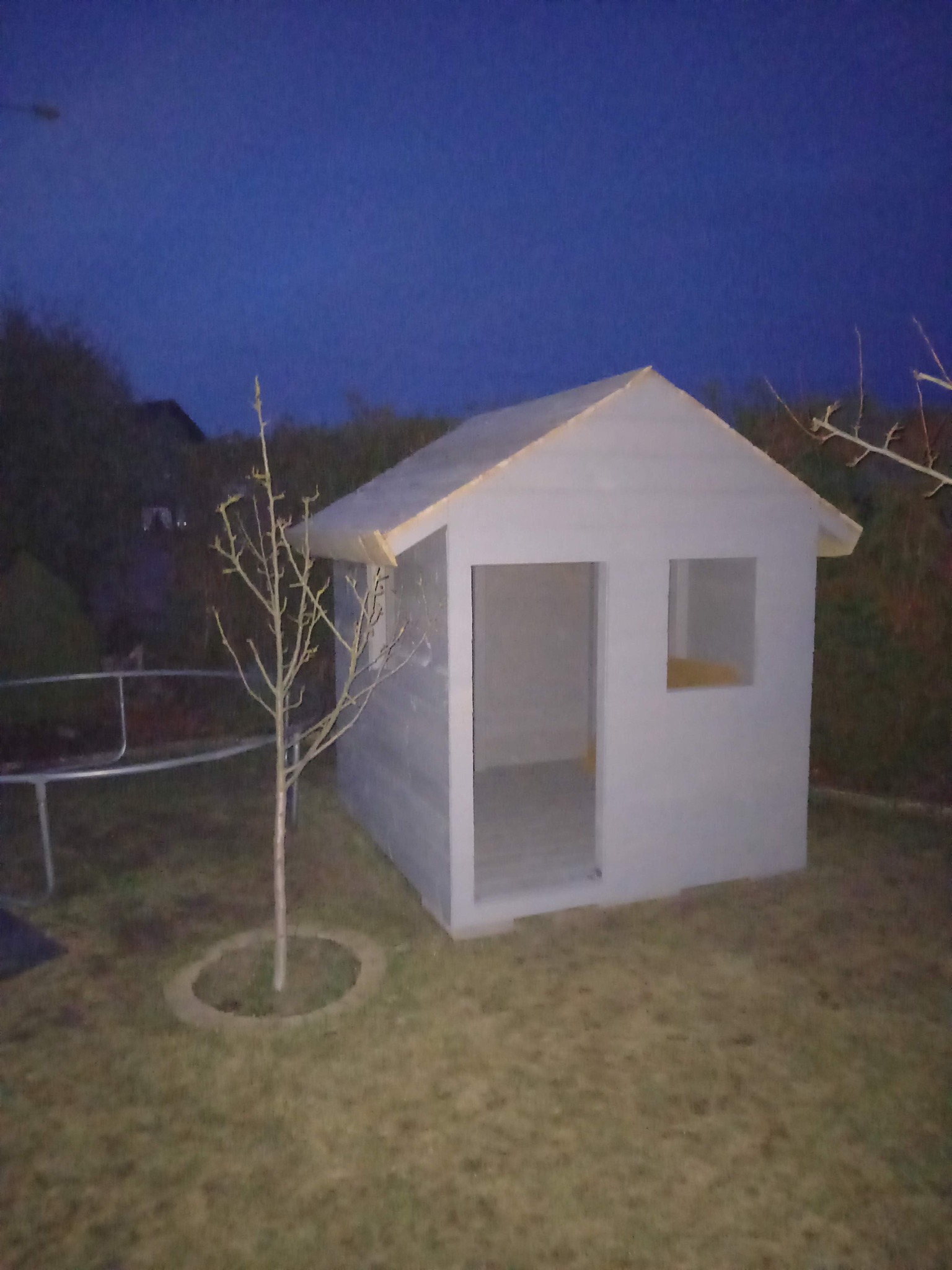 Mała, pomalowana na szaro drewniana altana z dwoma otworami okiennymi, stojąca na trawniku obok młodego drzewka. Nocne oświetlenie.