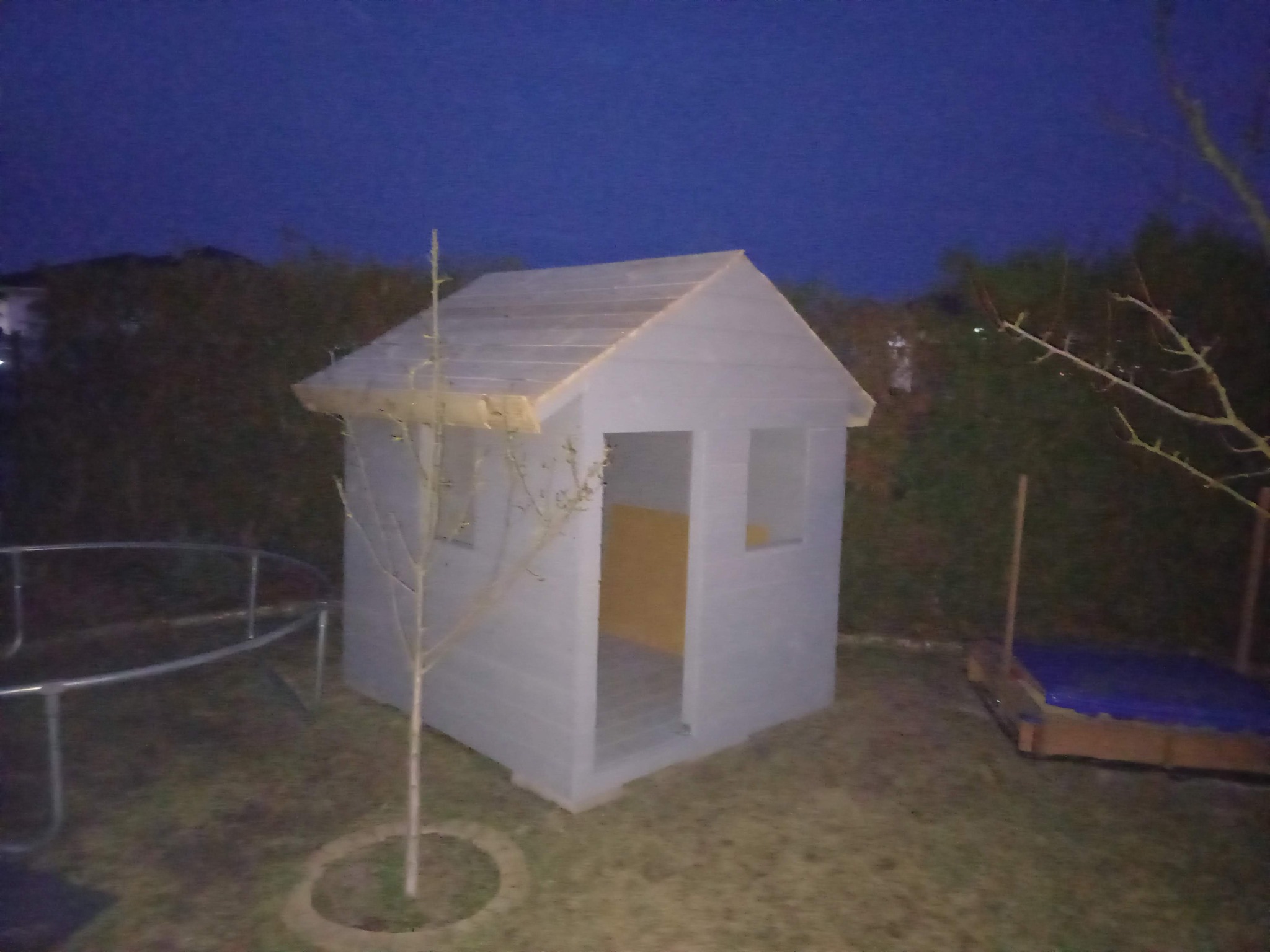 Mały, szary domek zabaw dla dzieci z drewnianą konstrukcją dachu, umieszczony w ogrodzie, widoczny przy zmierzchu. Obok trampolina i piaskownica.