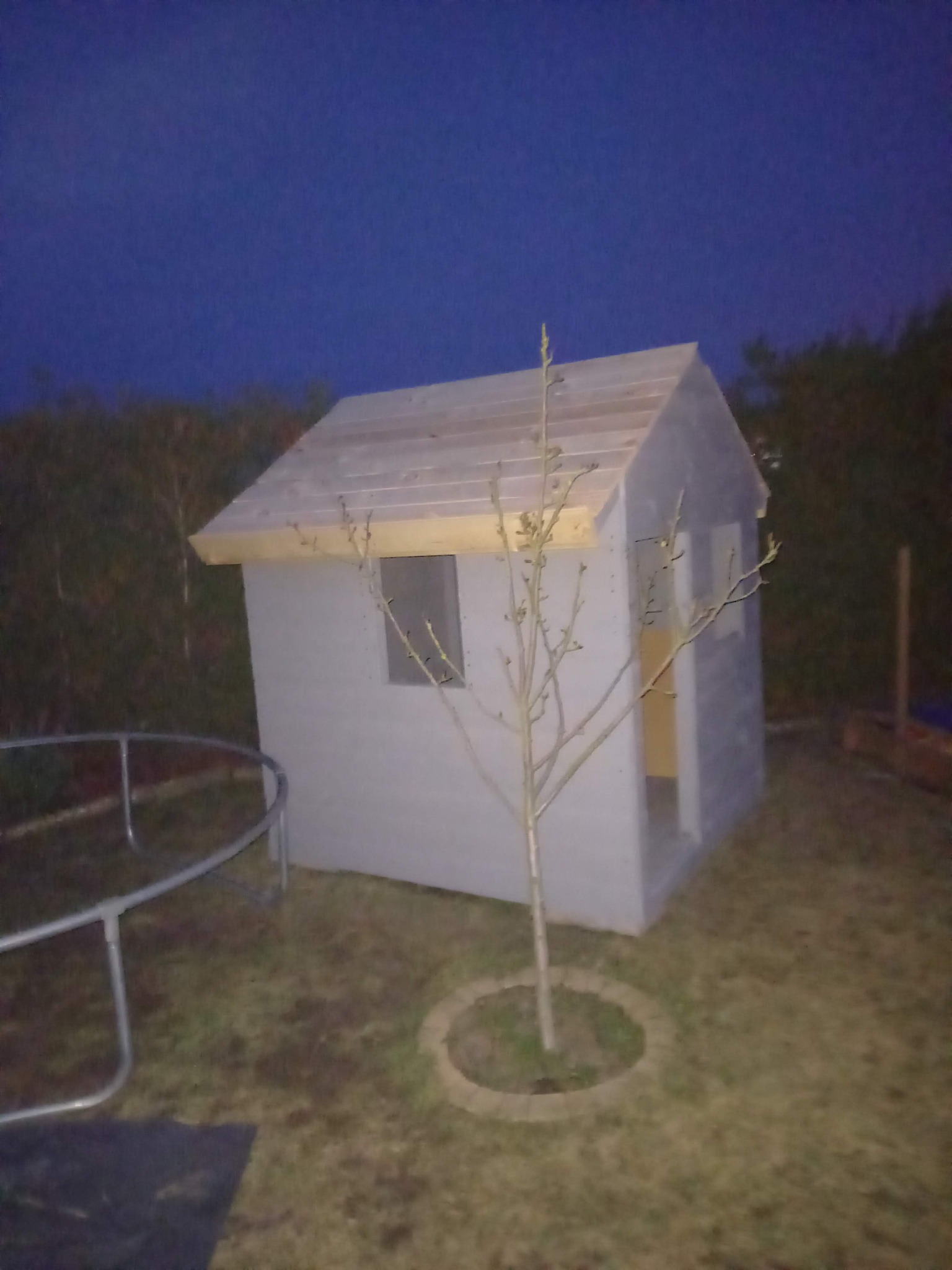 Mały, szary domek zabaw dla dzieci z drewnianym dachem, umieszczony na trawniku obok trampoliny, sfotografowany o zmierzchu. Przed domkiem rośnie małe drzewko.