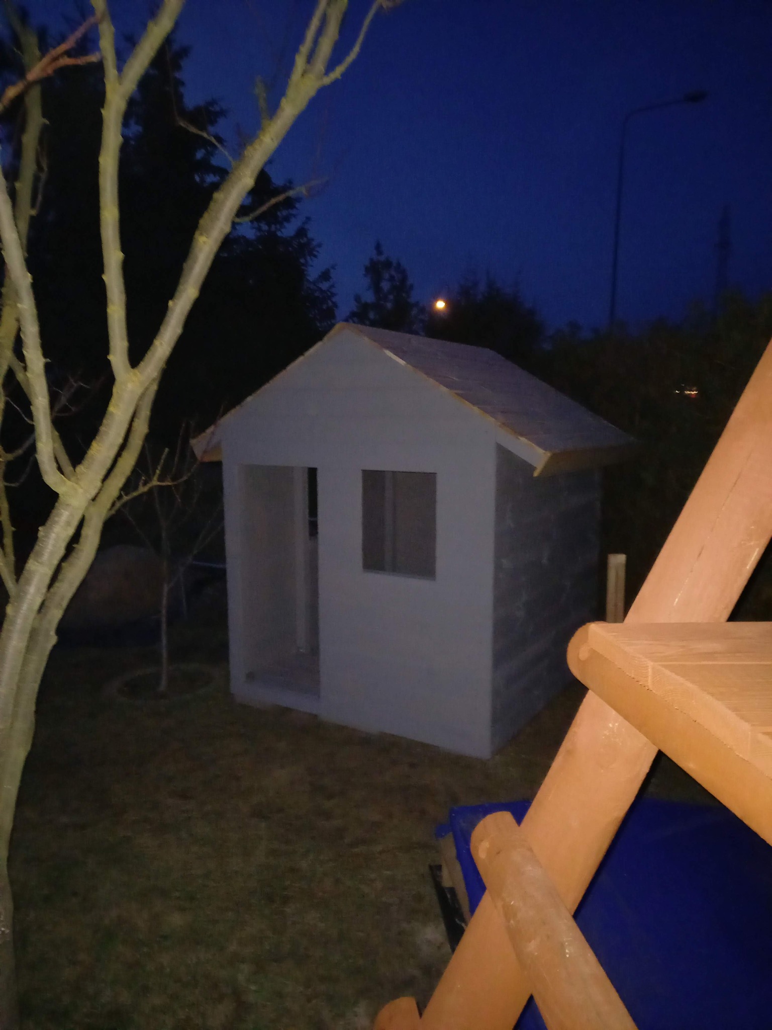 Mały, szary domek zabaw dla dzieci z drewnianym placem zabaw w tle, sfotografowany o zmierzchu. Widoczny fragment drzewa po lewej stronie kadru.