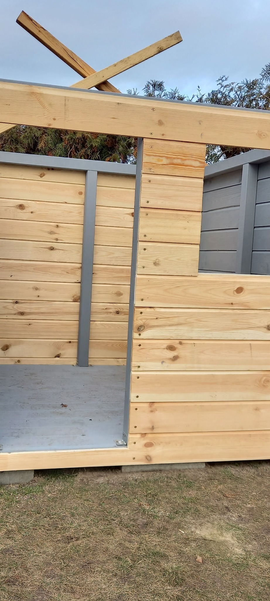 Surowa konstrukcja altany z jasnego drewna i szarymi elementami, widoczne łączenia śrubami, częściowo zadaszona, na tle zieleni i pochmurnego nieba.