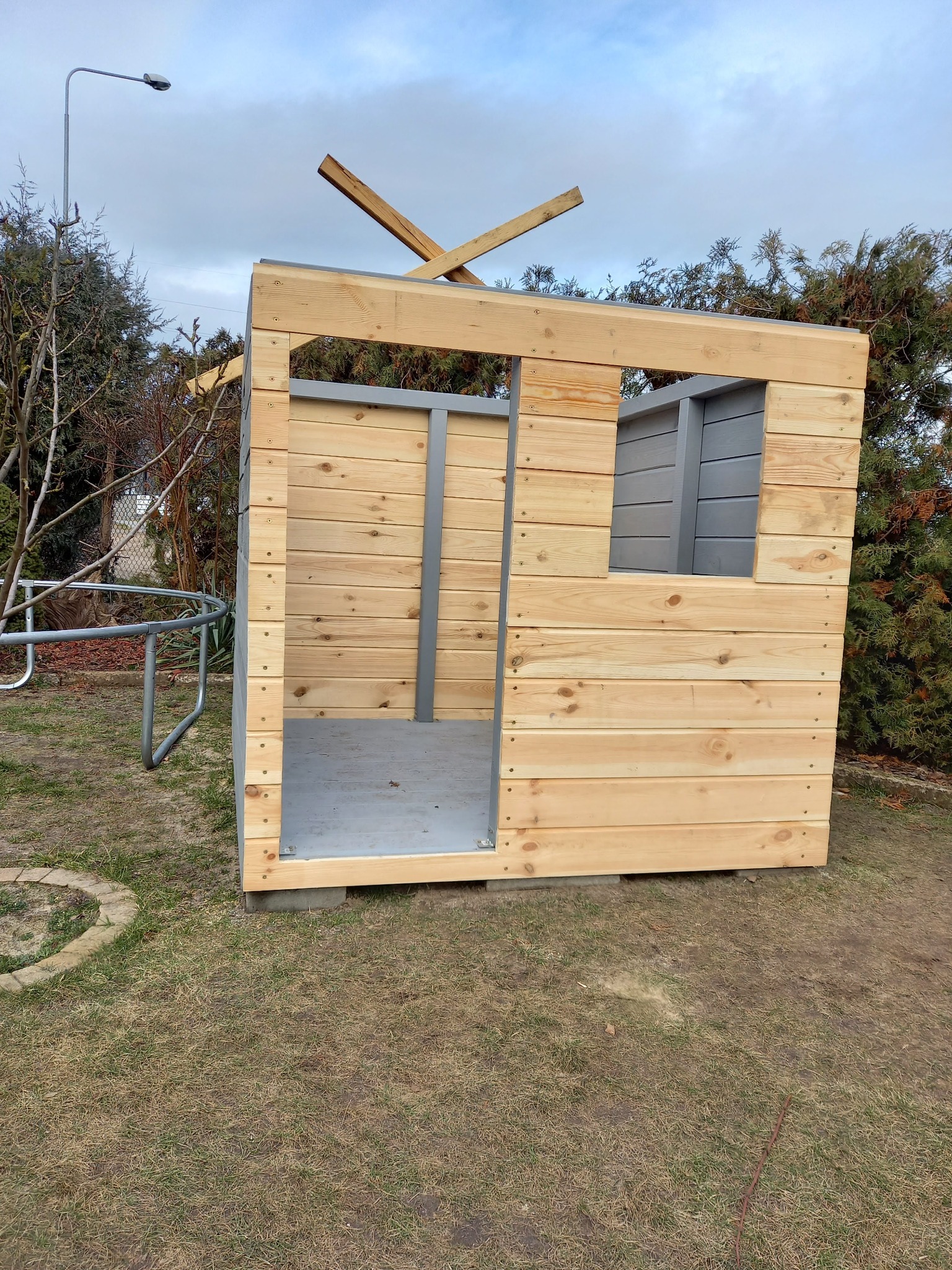 Niedokończona drewniana konstrukcja domku dla dzieci z szarą ramą okienną i częściowo zamontowanym dachem, stojąca na trawniku w ogrodzie.